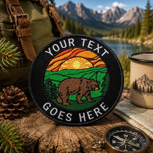 May include: A circular embroidered patch featuring a brown bear walking through a green forest landscape under a yellow and orange sunset. The black border contains the white embroidered text YOUR TEXT GOES HERE in a bold, sans-serif font.