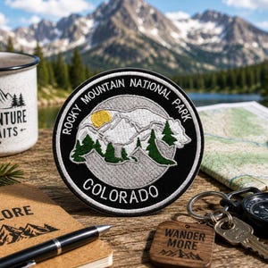 May include: A circular black and white patch with the text "Rocky Mountain National Park Colorado" encircling a mountain and bear design. A wooden keychain with the words "Wander More" is also visible.