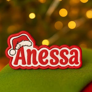 May include: A red and white embroidered patch with the name "Anessa" in a festive font, topped with a Santa hat design. The patch is set against a green surface, suggesting a Christmas theme.