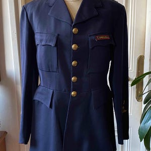 Peut inclure: Veste militaire vintage bleu marine avec des boutons dorés et un écusson "COMMANDOS". La veste a deux poches de poitrine et deux poches inférieures. La veste est présentée sur un mannequin.