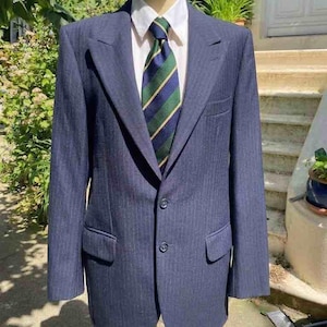 Peut inclure: Veste de costume bleu marine avec chemise blanche à col et cravate rayée verte, bleue et dorée. La veste a une fermeture à deux boutons et des poches avant. Le costume est présenté sur un mannequin en bois.