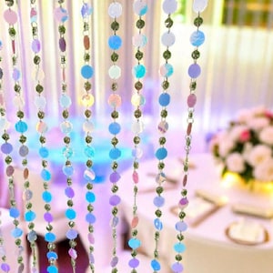 May include: Iridescent beaded curtain with round, colorful sequins in shades of blue, purple, and pink. The curtain is strung on a green thread and is likely used for decorative purposes at an event.