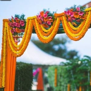May include: A wedding arch decorated with yellow and orange marigold flower garlands and orange fabric drapes.