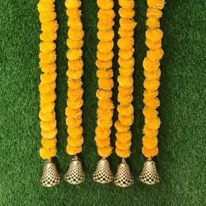 May include: Six strands of yellow marigold flower garlands with gold bells at the bottom. The garlands are hanging against a green background.