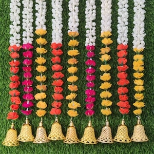 May include: Ten white flower garlands with colorful flower accents and gold bells at the bottom. The garlands are hanging against a green background.