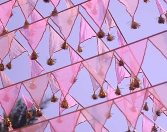 Könnte beinhalten: Dekorative Girlande aus rosa und weißen Wimpelfahnen vor blauem Himmel. Jede Fahne ist mit einer goldenen Quaste verziert, was eine festliche und elegante Ästhetik erzeugt. Die Anordnung deutet auf eine Feier oder ein Ereignis hin.