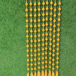 May include: A decorative yellow beaded curtain with gold accents and tassels. The curtain is made of rows of yellow beads and small gold beads, with a tassel at the bottom of each row. The background is green.