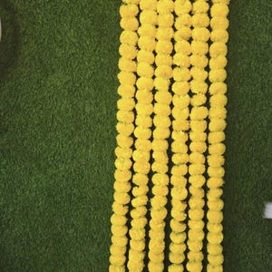 May include: Image showing multiple strands of bright yellow marigold flower garlands against a green backdrop. The garlands are densely packed with round, fluffy flowers, creating a vibrant and festive display.