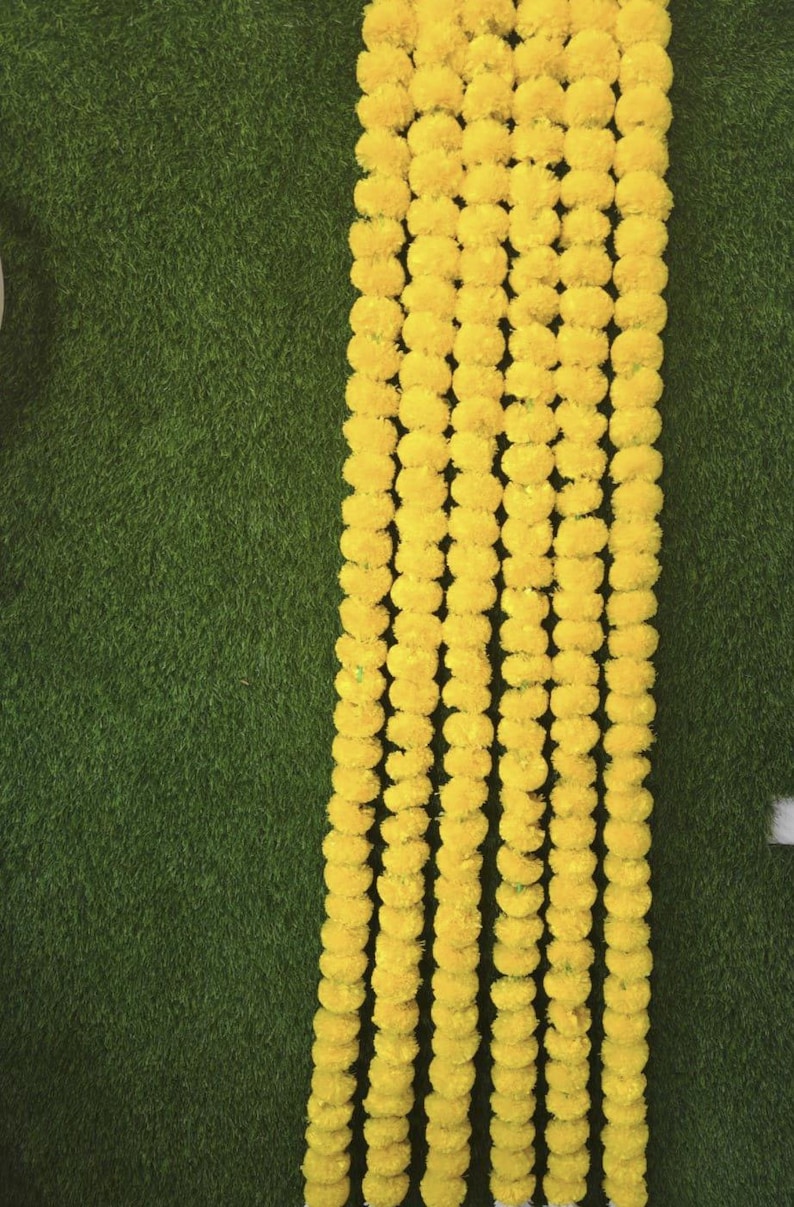 May include: Image showing multiple strands of bright yellow marigold flower garlands against a green backdrop. The garlands are densely packed with round, fluffy flowers, creating a vibrant and festive display.
