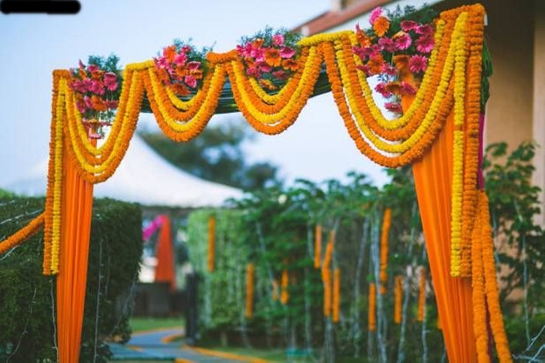 May include: A wedding arch decorated with yellow and orange marigold flower garlands and orange fabric drapes.