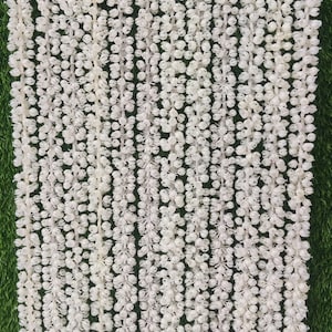 Pode incluir: Guirlanda floral branca pendurada contra um fundo verde. A guirlanda é feita de pequenas flores brancas redondas, amarradas em fileiras verticais, criando uma aparência densa e texturizada. Ideal para decorações de eventos.