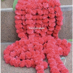 May include: A bright red floral garland, possibly made of marigolds, displayed in a cascading design. The garland is densely packed with small, round flowers, creating a textured appearance. Green leaves are visible at the top, resting on a stone surface.