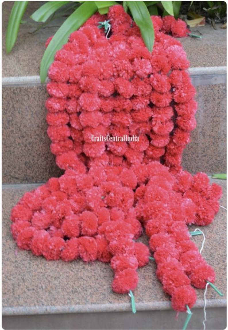 May include: A bright red floral garland, possibly made of marigolds, displayed in a cascading design. The garland is densely packed with small, round flowers, creating a textured appearance. Green leaves are visible at the top, resting on a stone surface.