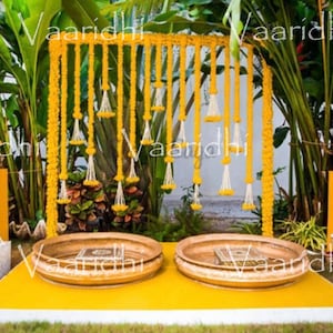 May include: A traditional Indian wedding ceremony setup featuring a floral garland backdrop made of yellow marigolds, two golden bowls, and a yellow and white platform.