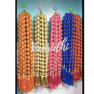 Pode incluir: Cinco fios de contas de tecido coloridas com borlas penduradas em ganchos. As contas são laranja, dourado, rosa, azul e vermelho. Os fios têm todos o mesmo comprimento e estão pendurados em fila. O texto "Vaaridhi" é visível na imagem.