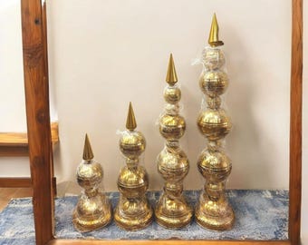 Puede incluir: Cuatro soportes decorativos de metal dorado con una parte superior puntiaguda y varios niveles redondos. Los soportes están dispuestos en orden de tamaño, con el soporte más grande a la izquierda y el más pequeño a la derecha.