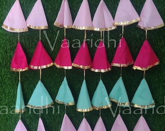 May include: A garland of pink, teal and magenta fabric cones with gold trim. The cones are strung together on a string and hang from a wall. The garland is decorated with the word "Vaaridhi" in gold lettering.