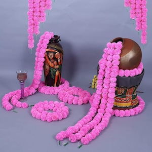 May include: An arrangement of pink floral garlands draped around two decorative vases. The vases are brown and decorated with painted figures. A wine glass and a circular garland are also present. The garlands hang from above.