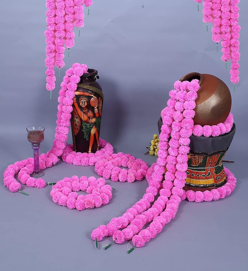 May include: An arrangement of pink floral garlands draped around two decorative vases. The vases are brown and decorated with painted figures. A wine glass and a circular garland are also present. The garlands hang from above.