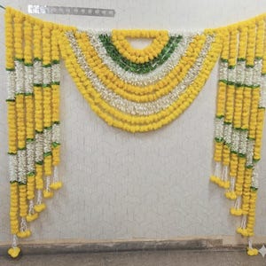 Pode incluir: Um adorno decorativo para porta com fios verticais alternados de flores de calêndula amarelas e flores brancas semelhantes a jasmim. A seção central forma uma guirlanda semicircular em camadas com detalhes verdes, enquanto as laterais pendem.