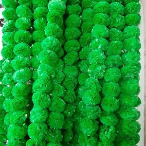 May include: A close-up of multiple strands of bright green, spherical artificial flower garlands. The garlands are densely packed, creating a textured, vibrant green backdrop. The flowers appear to be made of a soft, possibly fabric-like material.