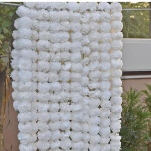 May include: A vertical display of white floral garlands, possibly for decoration. The garlands are made up of many tightly packed, round, fluffy white flowers. The background shows greenery and a building, suggesting an outdoor setting.