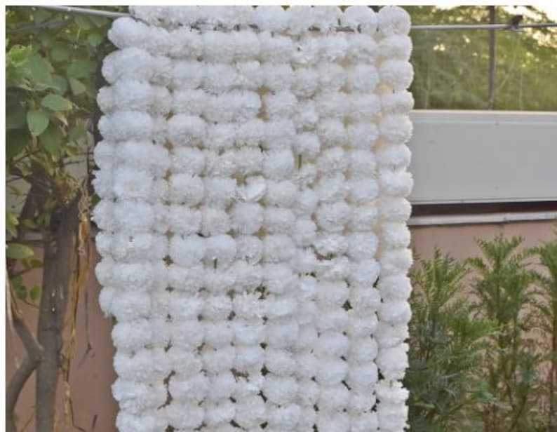 May include: A vertical display of white floral garlands, possibly for decoration. The garlands are made up of many tightly packed, round, fluffy white flowers. The background shows greenery and a building, suggesting an outdoor setting.