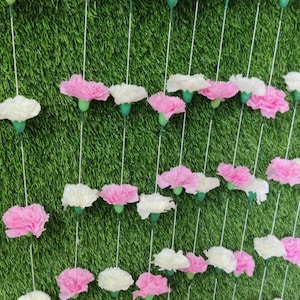 May include: A garland of pink and white carnations strung together with white thread. The garland is hanging against a green artificial grass backdrop.