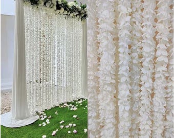 May include: A white floral backdrop for a wedding or event. The backdrop features cascading white flower garlands, a floral arrangement at the top, and white fabric draping. The backdrop is set on a green lawn with scattered flower petals.