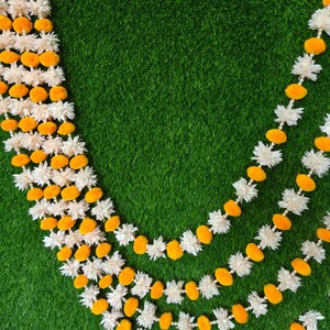 Pode incluir: Close-up de grinaldas decorativas com elementos florais brancos e laranja alternados. As grinaldas são dispostas sobre um fundo verde, criando um contraste vibrante. O design floral é detalhado e simétrico, sugerindo um estilo artesanal.