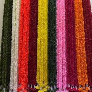 Puede incluir: Una colección de coloridas guirnaldas de flores artificiales colgadas verticalmente. Las guirnaldas son de varios colores, incluyendo verde, blanco, naranja, rojo, amarillo, rosa y burdeos. Cada guirnalda tiene un grupo floral decorativo en la parte inferior.