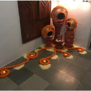 May include: A decorative arrangement of orange flower garlands and candles on a tiled floor.  The garlands are arranged in a swirling pattern, and the candles are placed in the center of each swirl.  There are also several terracotta pots with lids, decorated with orange flowers, in the background.