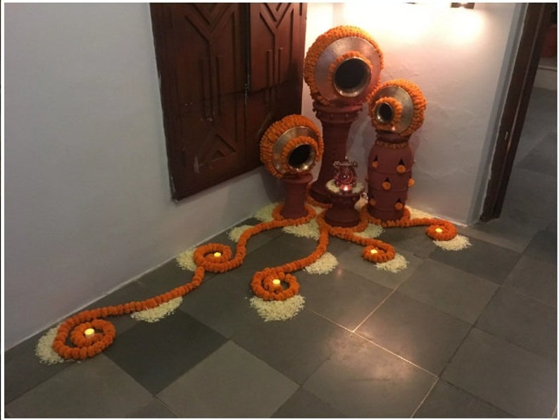May include: A decorative arrangement of orange flower garlands and candles on a tiled floor.  The garlands are arranged in a swirling pattern, and the candles are placed in the center of each swirl.  There are also several terracotta pots with lids, decorated with orange flowers, in the background.