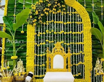 May include: A traditional Indian wedding ceremony backdrop featuring a yellow flower garland archway and a white and gold chair. The backdrop is made of green banana leaves.