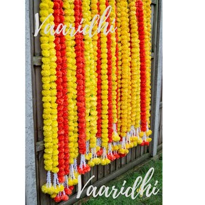 May include: A garland of yellow and orange marigold flowers, arranged in alternating rows, hangs against a wooden fence. The garland is labeled "Vaaridhi".