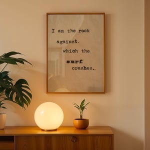 May include: A white globe lamp with a wooden base sits on a wooden table next to a potted plant. A framed print with the text "I am the rock against which the surf crashes." hangs on a beige wall.