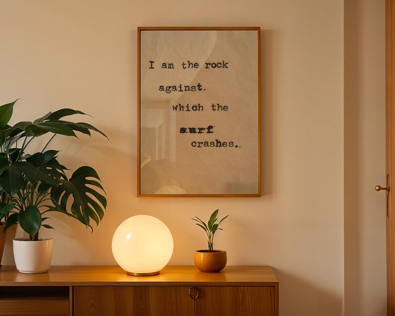 May include: A white globe lamp with a wooden base sits on a wooden table next to a potted plant. A framed print with the text "I am the rock against which the surf crashes." hangs on a beige wall.