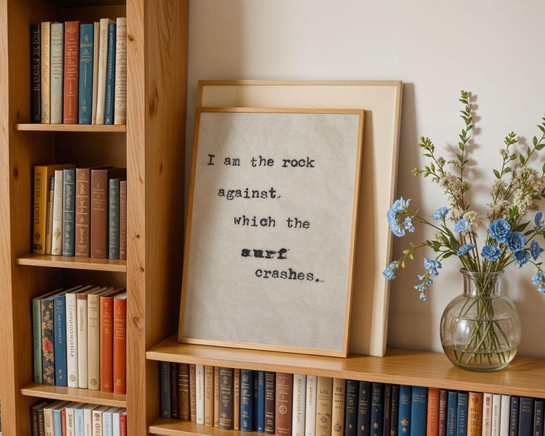 May include: A framed print with a quote about the ocean: "I am the rock against which the surf crashes..." The print is hanging on a wooden shelf with books and a vase of blue flowers.