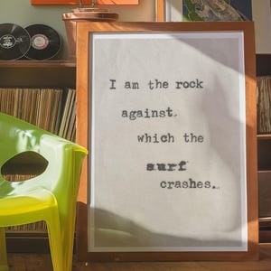 May include: A framed print with a quote in black typewriter font on a beige background. The quote reads "I am the rock against which the surf crashes..."