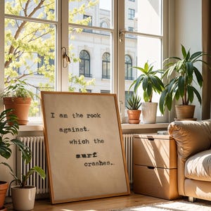 May include: A framed print with a quote about being the rock against which the surf crashes. The print is hanging on a wall in a living room with a couch and plants.