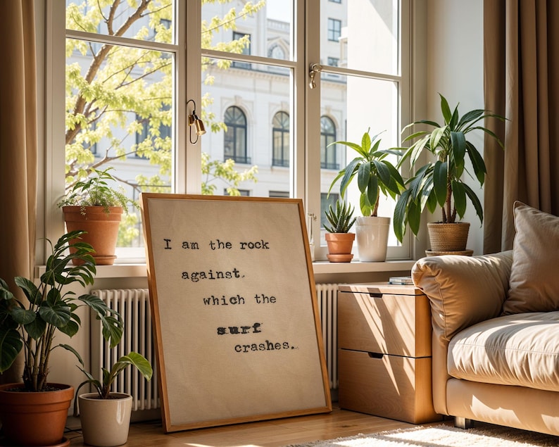 May include: A framed print with a quote about being the rock against which the surf crashes. The print is hanging on a wall in a living room with a couch and plants.