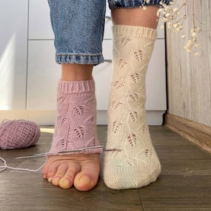 Könnte beinhalten: Ein Paar gestrickte Socken, eine rosa und eine weiße, mit einem Lochmuster. Die Socken werden mit Stricknadeln gearbeitet.