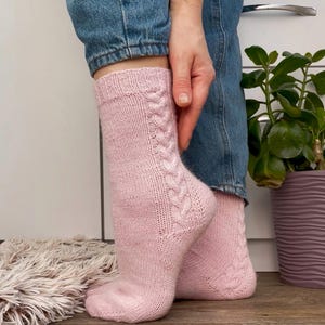 Puede incluir: Un par de calcetines de punto rosa claro con un diseño de trenzas en los laterales. Los calcetines se llevan con vaqueros azules. Los calcetines están hechos de un hilo suave y texturizado y parecen hechos a mano.