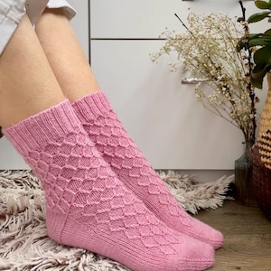 Peut inclure: Une paire de chaussettes tricotées rose clair présentant un motif en losanges texturé sur le pied et la cheville, avec un bord-côte en haut. Les chaussettes sont réalisées avec un point de tricot régulier et une finition lisse au niveau du talon.