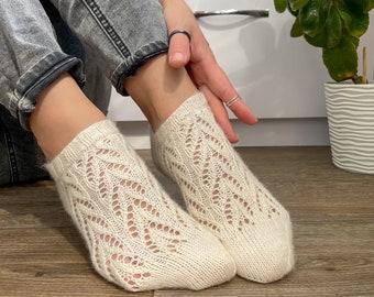 Könnte beinhalten: Ein Paar cremefarbene Socken mit offenem Gewebe und einem filigranen Muster. Die Socken bestehen aus einem weichen, gestrickten Material und werden an den Füßen einer Person getragen. Die Socken liegen auf einem Holzboden.