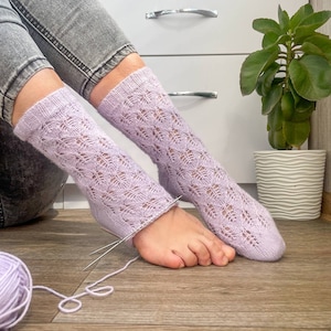 Peut inclure: Chaussettes tricotées ajourées lilas clair avec un motif de feuilles. Les chaussettes sont tricotées avec des aiguilles et du fil assorti. L'image montre les chaussettes sur les pieds d'une personne, avec une pelote de laine au premier plan.