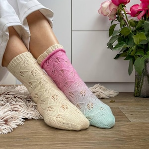 May include: Two knitted socks, one in cream and the other with a pink to blue gradient. The socks feature an openwork leaf design. A bouquet of pink roses is in the background. The socks are worn indoors.