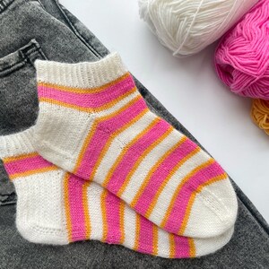 Puede incluir: Un par de calcetines blancos con rayas rosas y naranjas. Los calcetines están sobre un par de pantalones vaqueros grises.