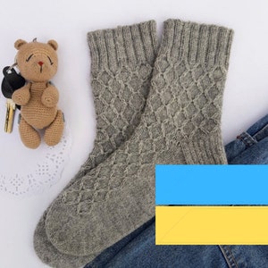 May include: A pair of grey knitted socks with a diamond pattern, beside a brown crocheted teddy bear keyring and a pair of blue jeans. The socks are made of a soft, warm material, ideal for cold weather.