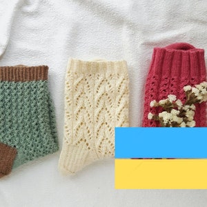 Könnte beinhalten: Drei Paar gestrickte Socken in verschiedenen Farben und Mustern werden auf einer weißen Oberfläche präsentiert. Die Socken umfassen ein grünes Paar mit brauner Ferse und Bündchen, ein cremefarbenes Paar mit Spitzendesign und ein rotes Paar mit kleinen weißen Blumen.